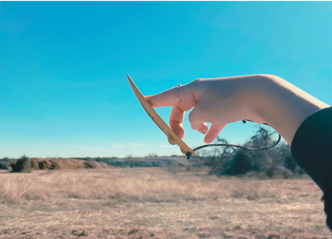 A woman's hand reaching out a car window with her pointer finger and middle finger on a wooden miniature surfboard, flying on the wind.