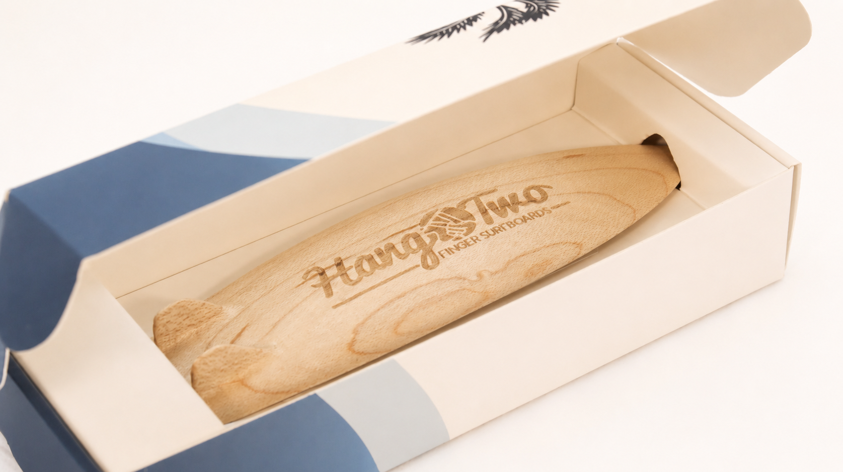 A wooden surfboard-shaped item with engraved text reading 'Hang Two Finger Surfboards' displayed inside an open off white-colored gift box with blue side panels, on a white background.