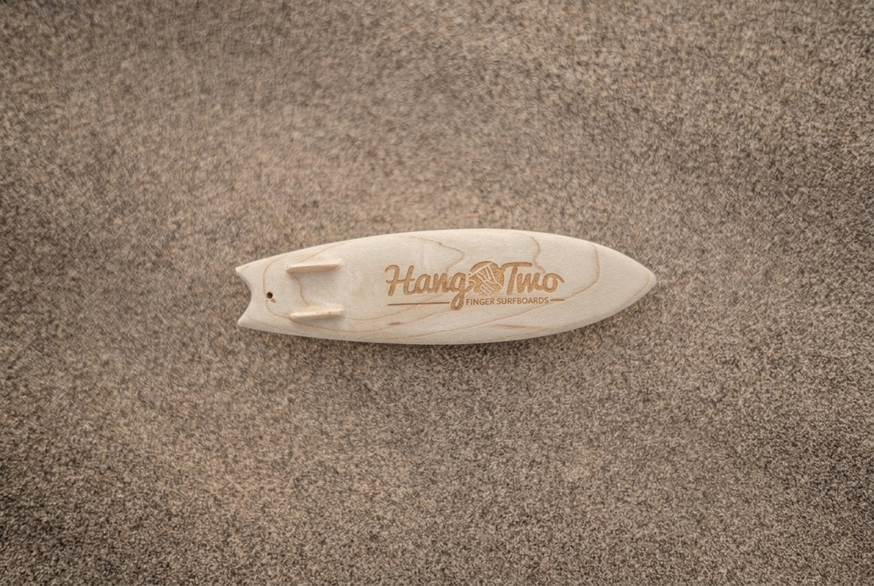 A small natural wood surfboard-shaped toy with the brand name 'Hang Two' engraved on top, placed on a sandy surface.