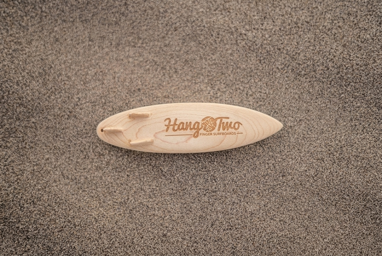 A small carved wooden surfboard made to fly out the car window with the brand name 'Hang Two' engraved, resting on sandy ground with soft shadows.
